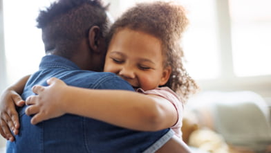 A child hugs her father while smiling in a sunny, well-lit room.