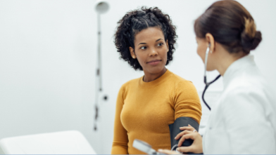 A woman in a mustard sweater attentively listens to a doctor with a stethoscope in a medical office.