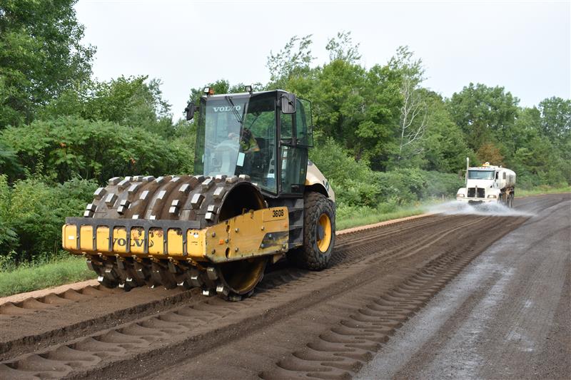 A heavy roller compacts brown soil on a road, with lush green trees in the background. A water truck sprays the soil, creating an industrious scene.