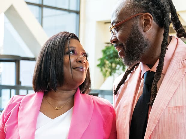 A couple dressed in pink blazers shares a warm, affectionate gaze in the atrium of Government Center, with large windows and greenery.