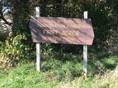 A brown sign next to a forest with text Reinvest in Minnesota First Acres in Hennepin County